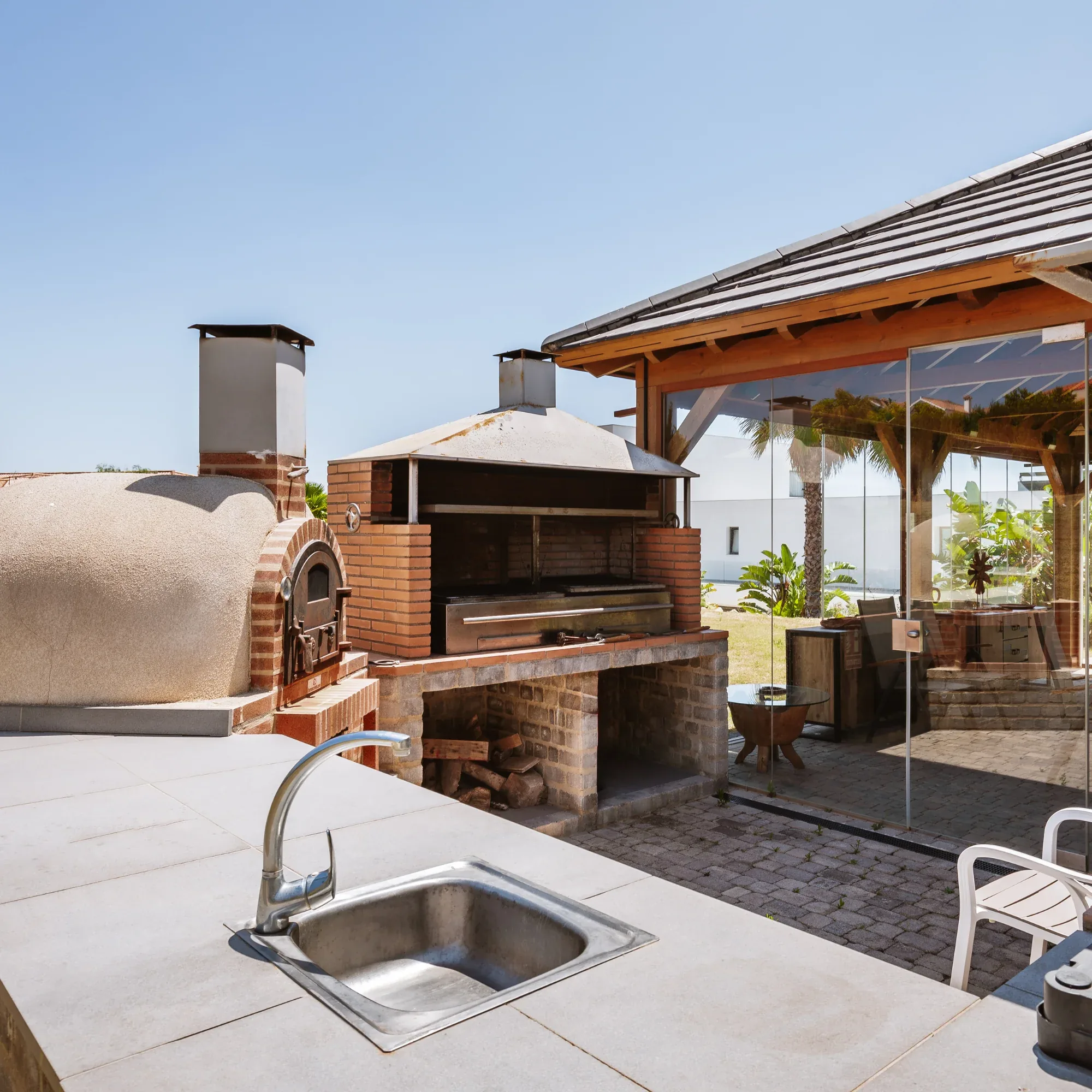 Outdoor kitchen with a brick pizza oven, barbecue grill and sink under a timber roofed pergola