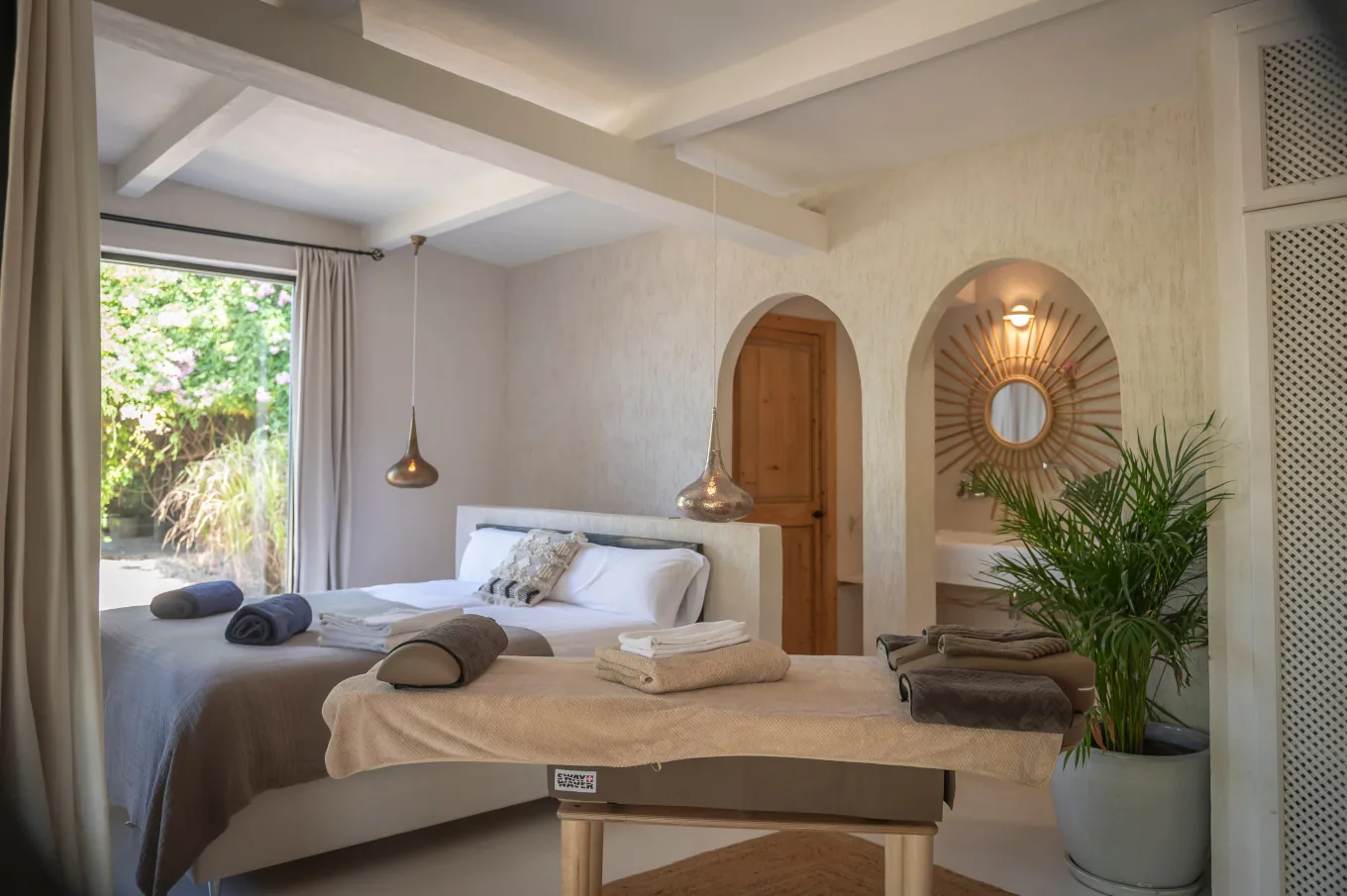 Bright villa bedroom with a white bed, arched doorway, sunburst mirror and a potted palm