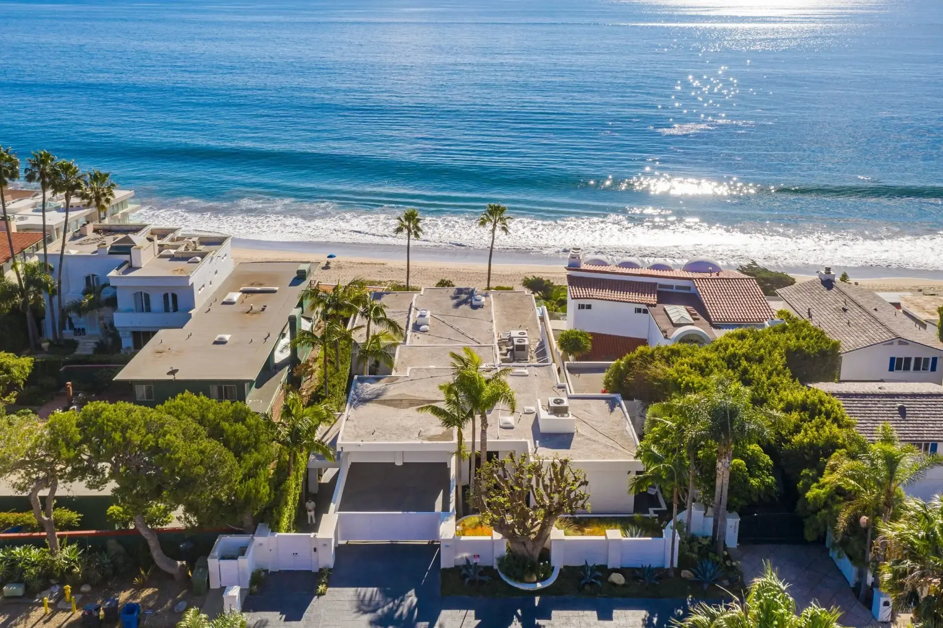 Aerial view over beachfront homes and palms toward a sunlit ocean with breaking waves