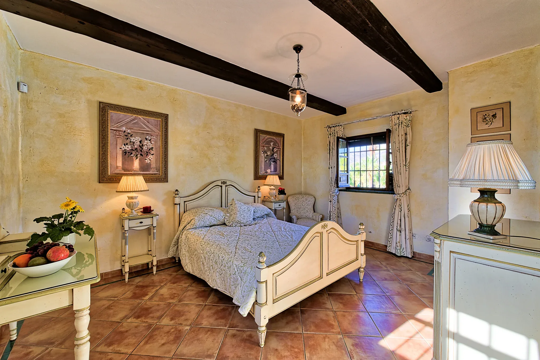 Client bedroom with dark wooden ceiling beams, a cream iron bed, terracotta floor and table lamps