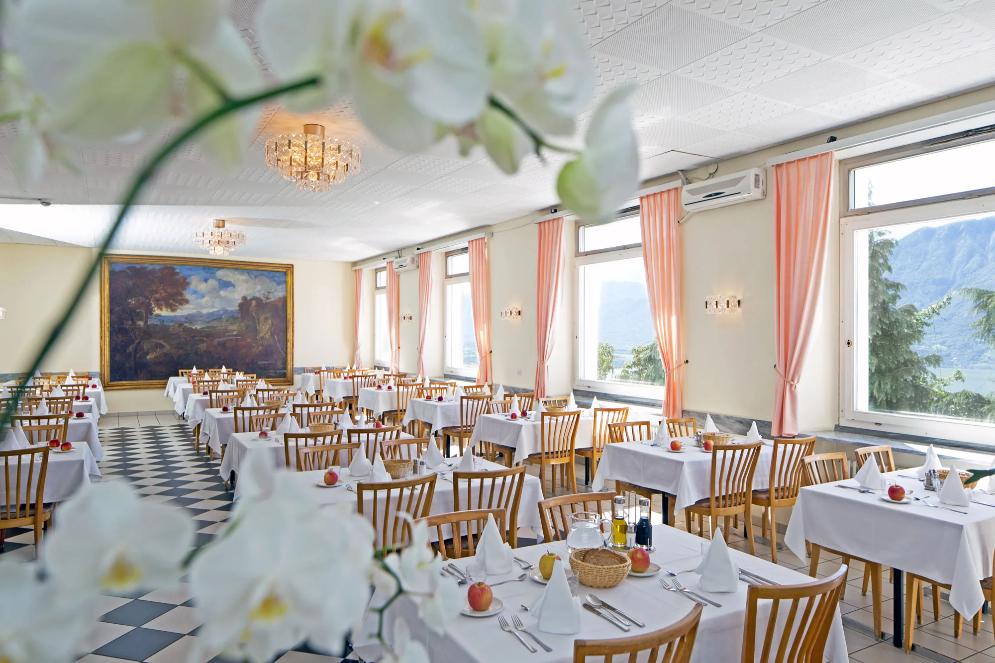 Large dining room with white tablecloths, rows of chairs and tall windows with peach curtains