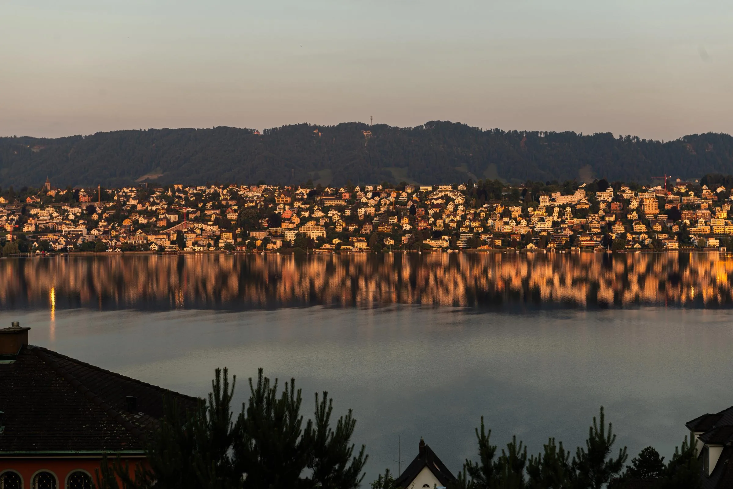 View over Lake Zurich at sunset with lit lakeside houses and wooded hills on the far shore