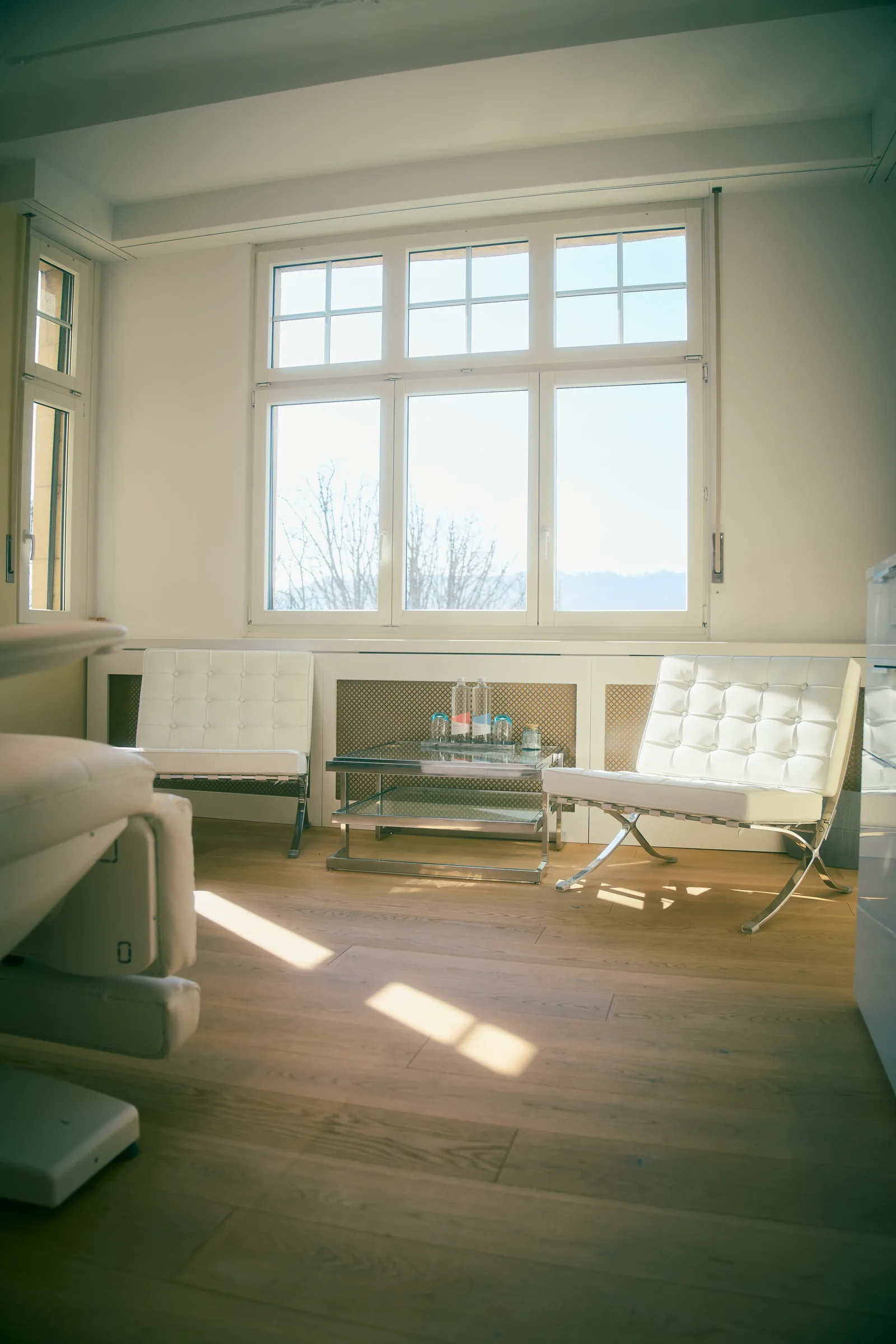 Medical treatment room with a padded couch, white armchairs and tall windows in daylight
