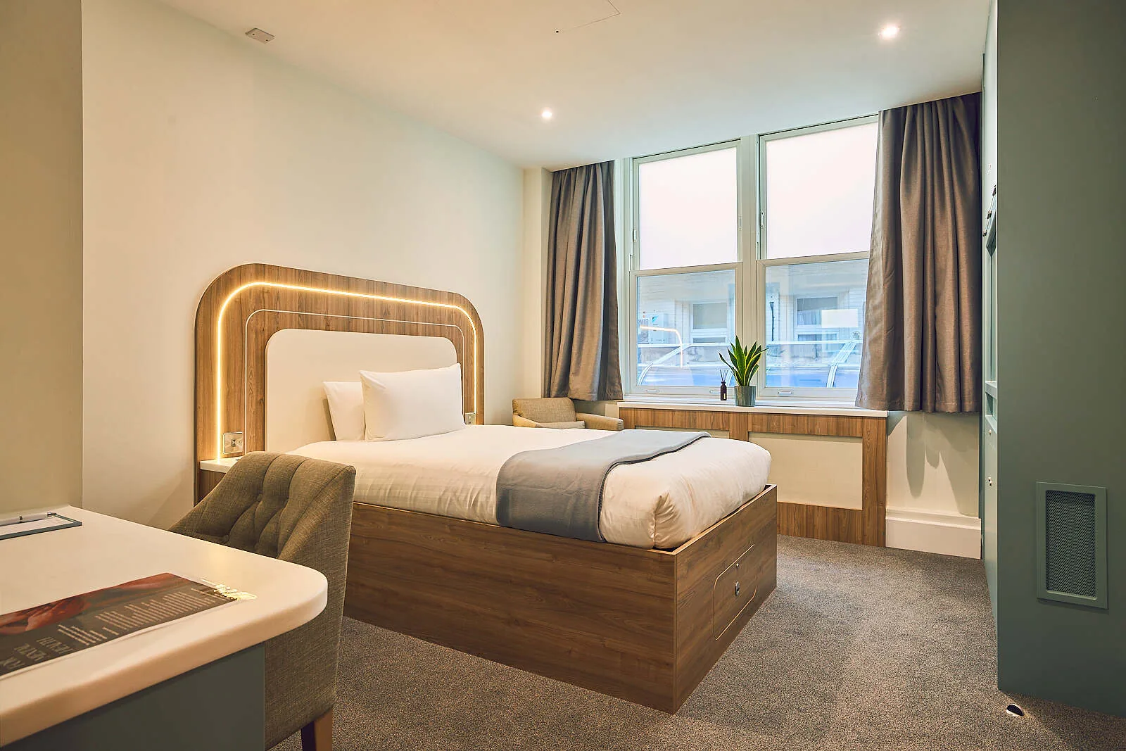 Single patient bedroom with a wooden bed frame, lit headboard, desk and a window with grey curtains