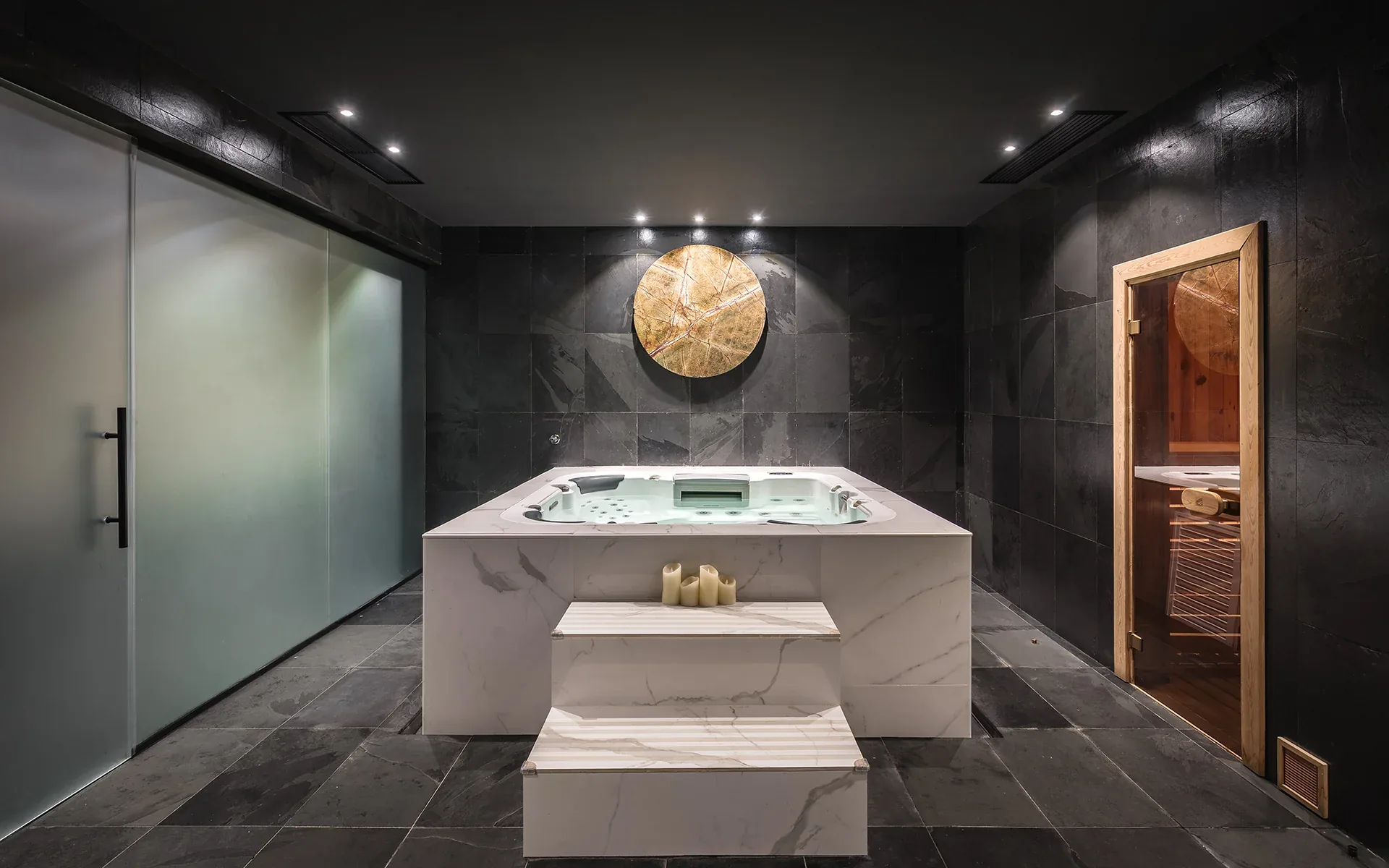Dark stone spa room with a white marble-clad jacuzzi, steps and a wooden sauna door
