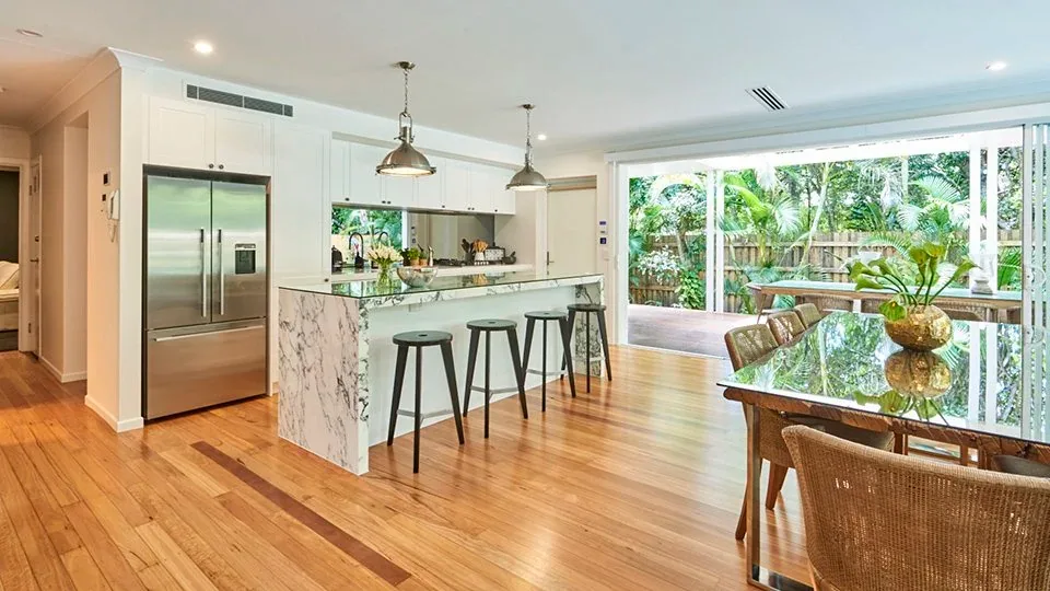 White kitchen with a marble island and bar stools beside a glass dining table and garden windows