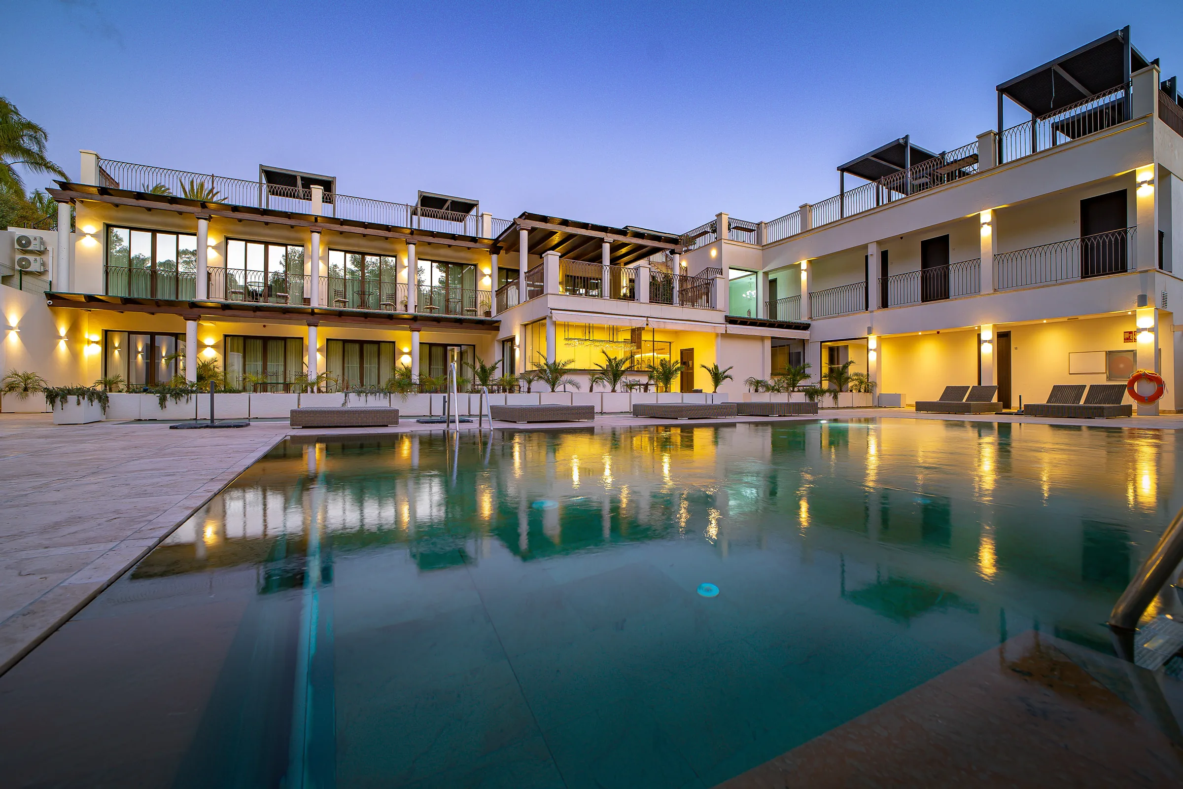 Three-storey white residence with lit balconies reflected in a long pool at dusk