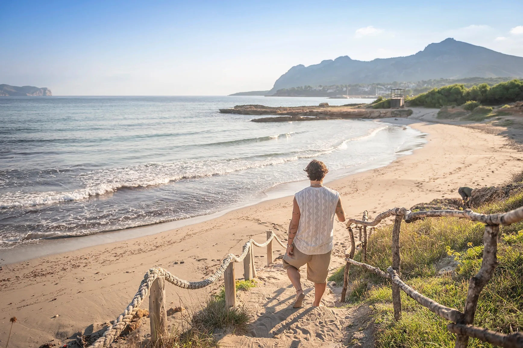 Person walking down a rope-lined path to a sandy beach with a headland and mountains behind