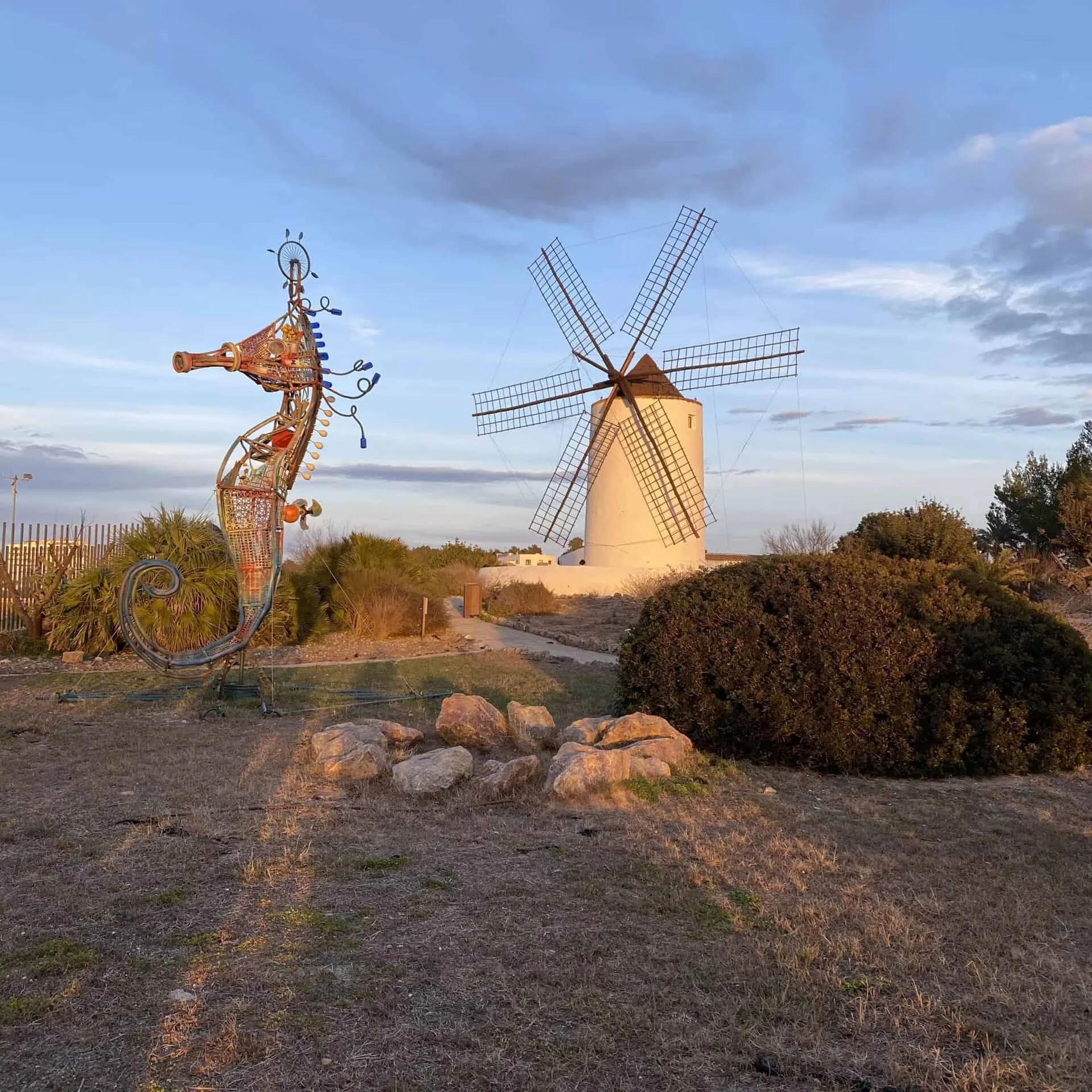 White stone windmill and a metal sculpture on a dry hilltop with shrubs under a blue sky