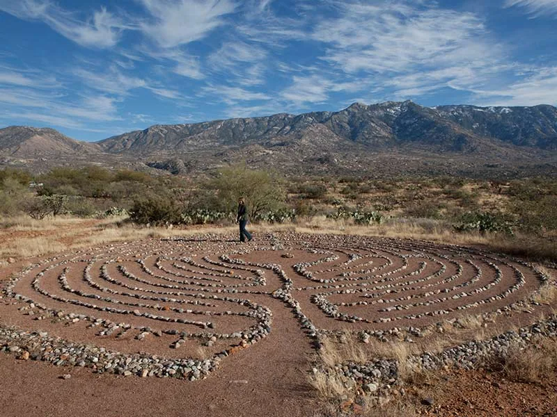 Stone labyrinth laid out on desert ground with scrub and a mountain range under a cloudy sky
