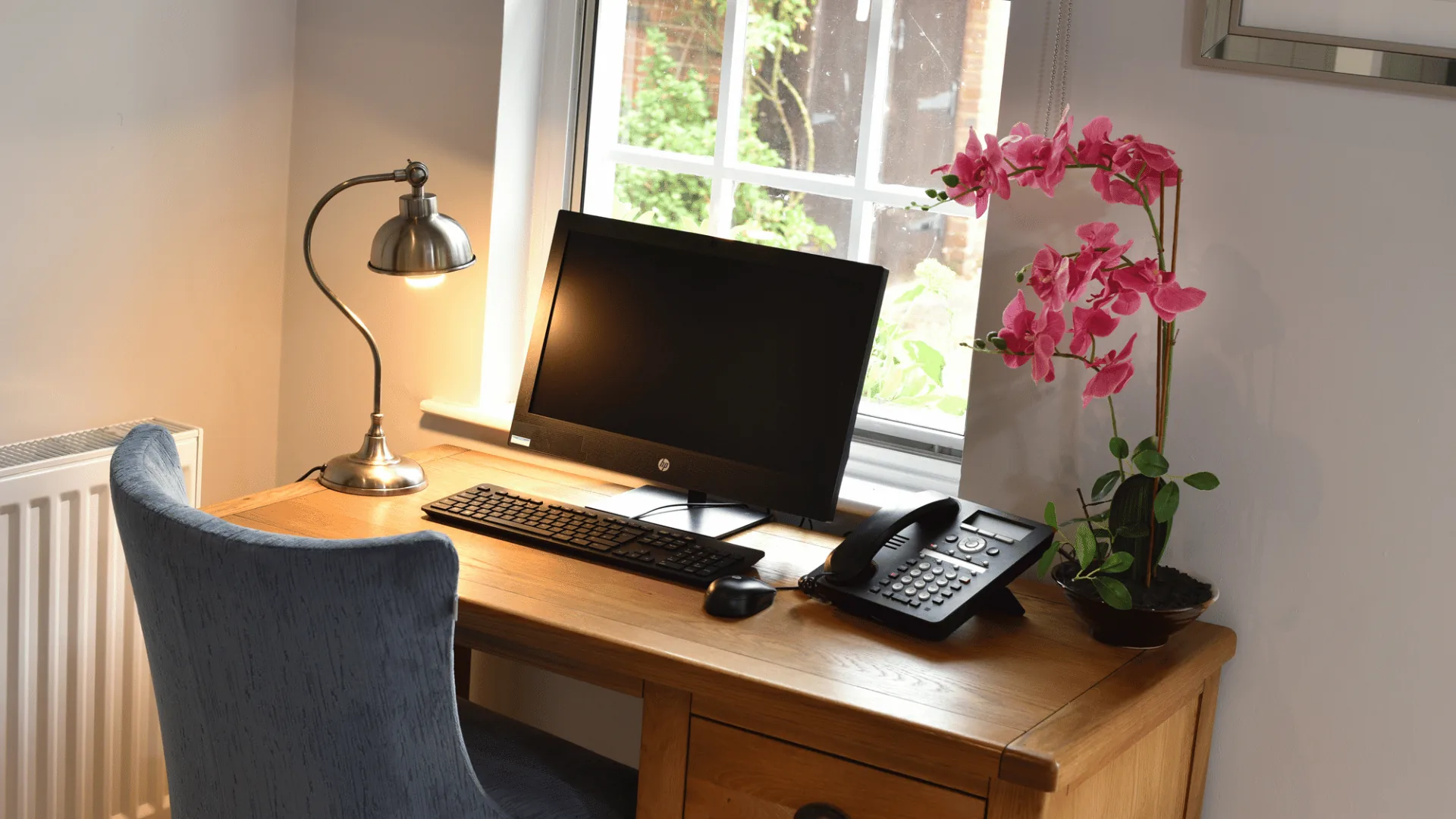 Wooden desk with a computer, desk lamp, telephone and pink orchid beside a leaded window