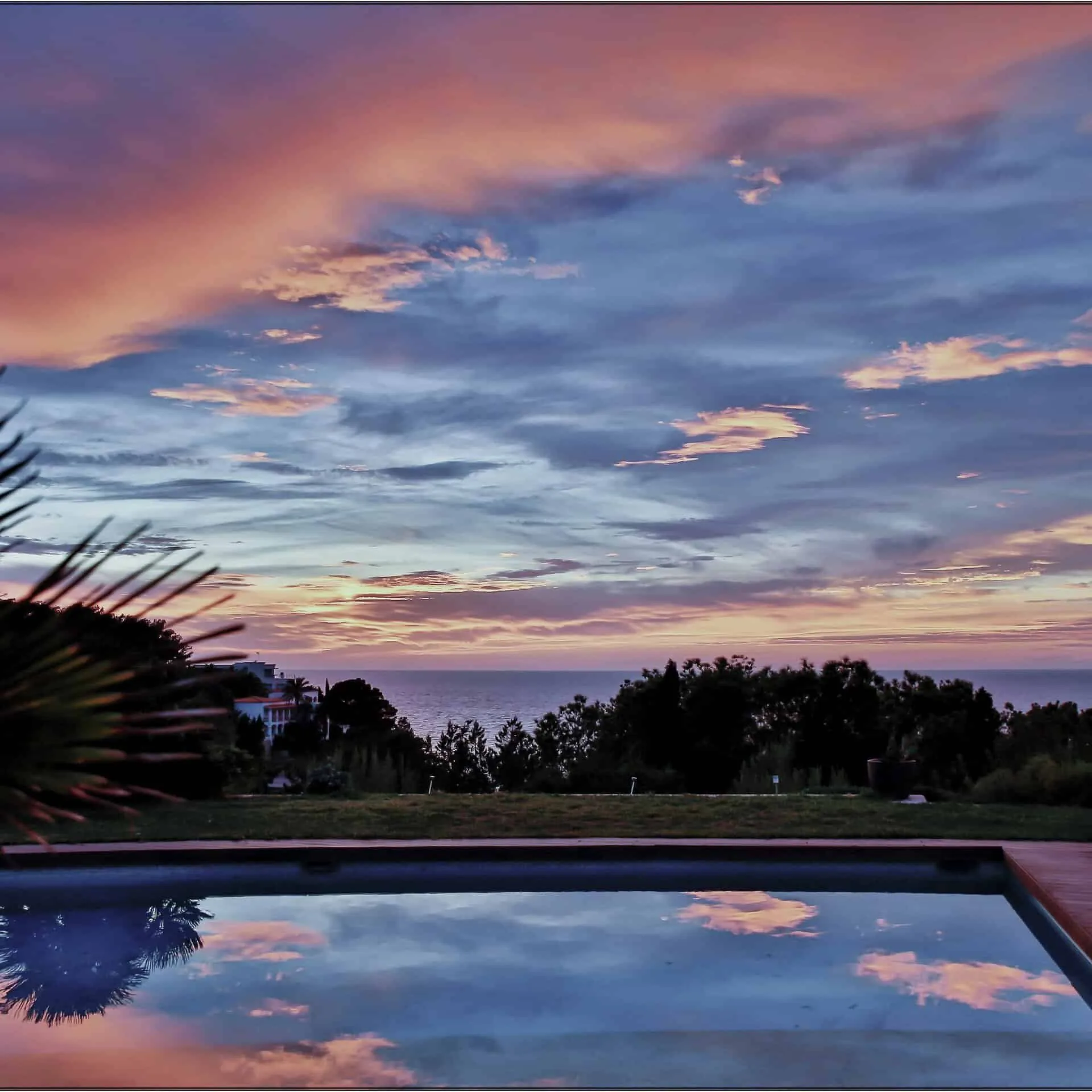 Pool reflecting a pink and blue sunset sky above dark trees and the sea, seen from an Ibiza residence
