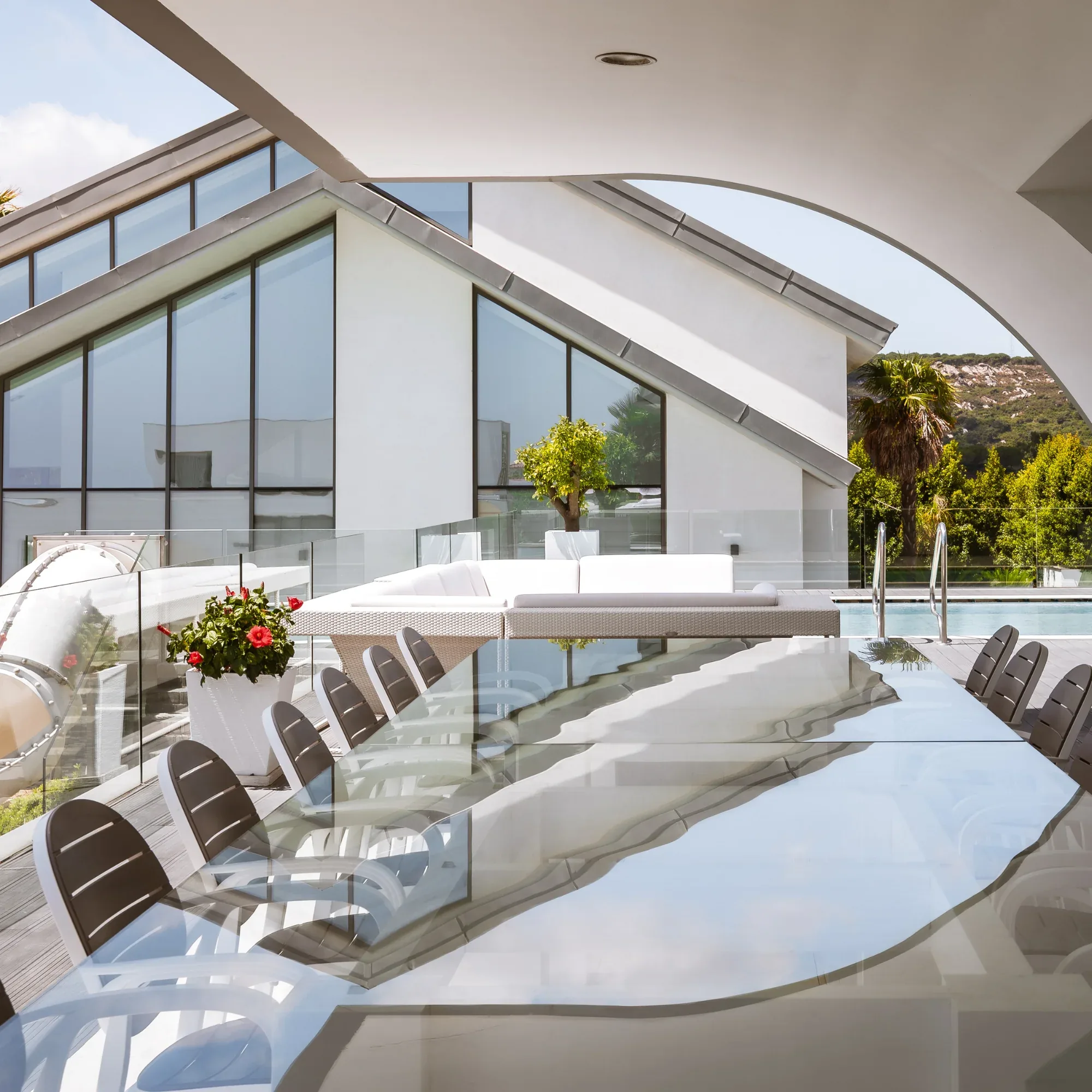 Long glass dining table with grey chairs on a terrace beside the pool and white villa buildings