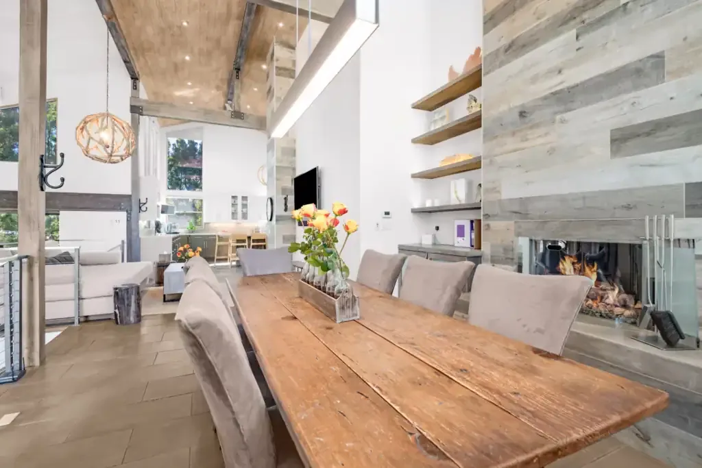 Long rustic wooden dining table with upholstered chairs beside a glass fireplace and open shelving