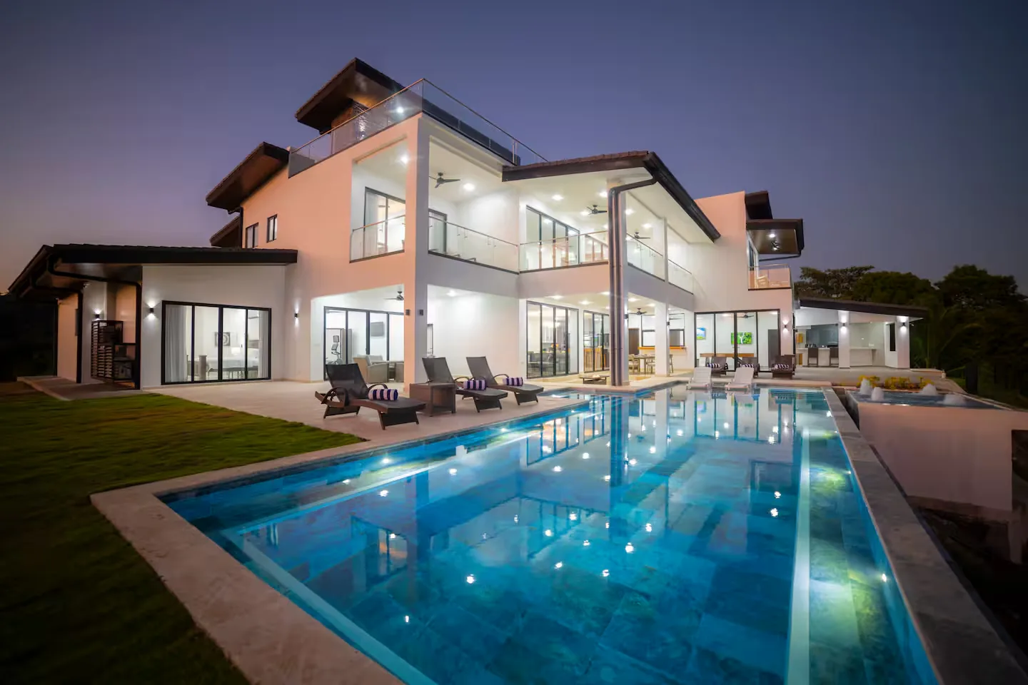 Modern multi-storey white villa at dusk with a lit infinity pool, sun loungers and a lawn