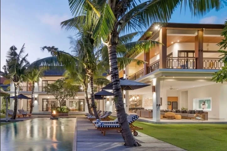 Two-storey villa with balconies behind a pool deck, sun loungers and tall palm trees in Bali
