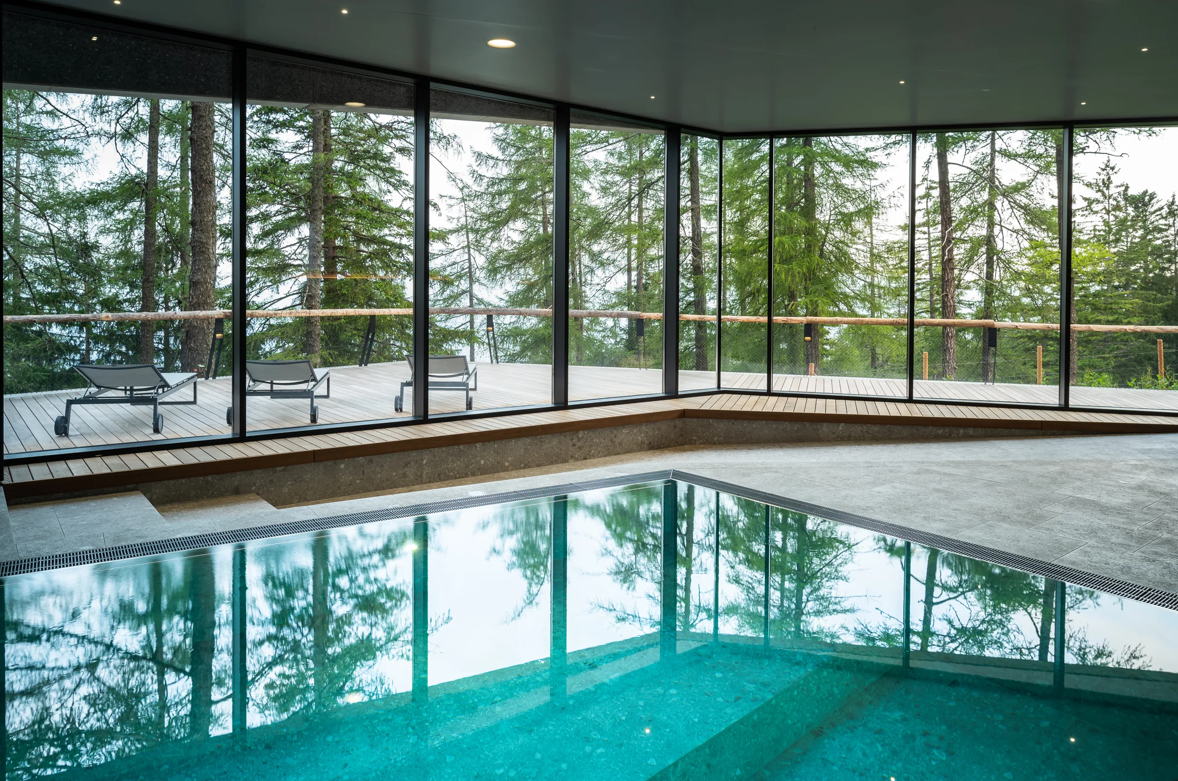 Indoor swimming pool with floor-to-ceiling glazing looking onto a terrace with loungers and tall conifers