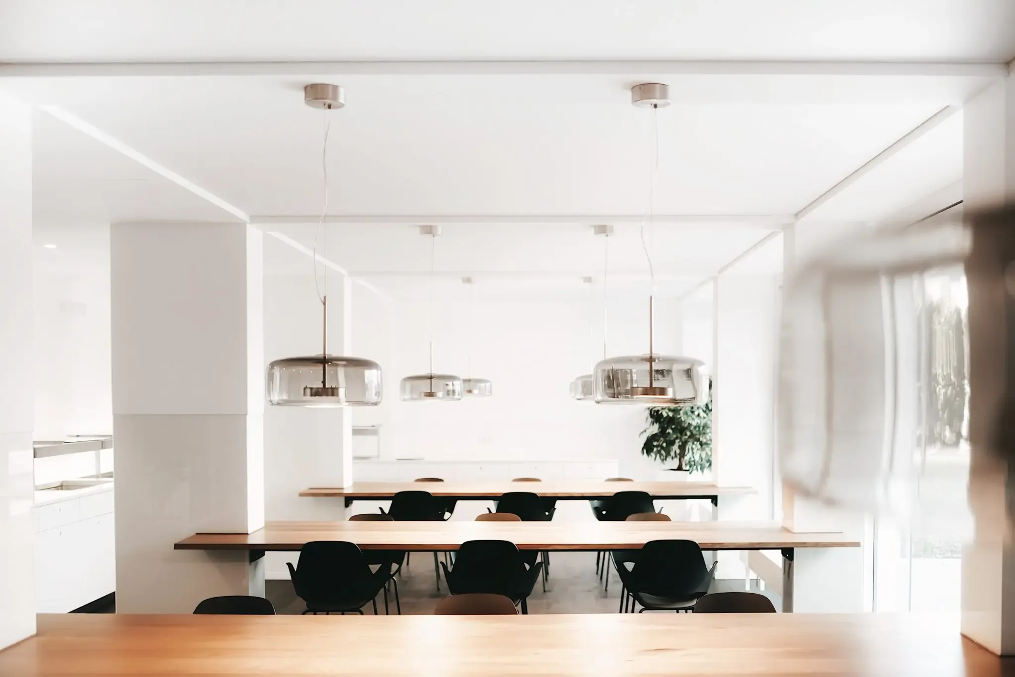 White dining room with long wooden tables, black chairs and glass pendant lamps hanging from the ceiling