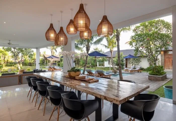 Long timber dining table with black chairs under rattan pendant lamps on a covered villa terrace in Bali