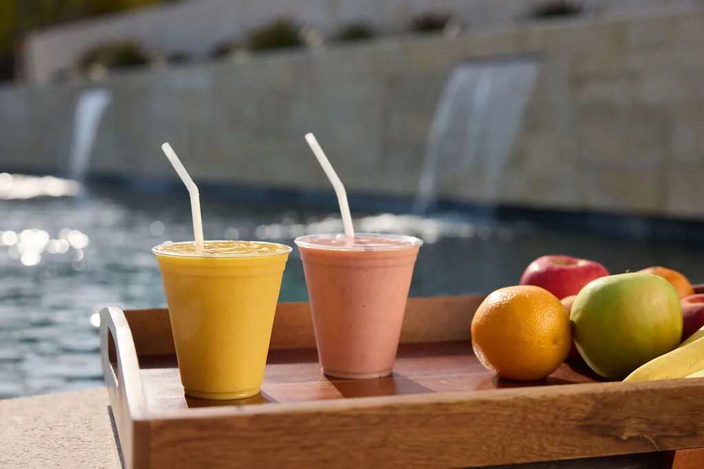 Tray with two smoothies and fresh fruit on the edge of a pool with a stone wall water feature