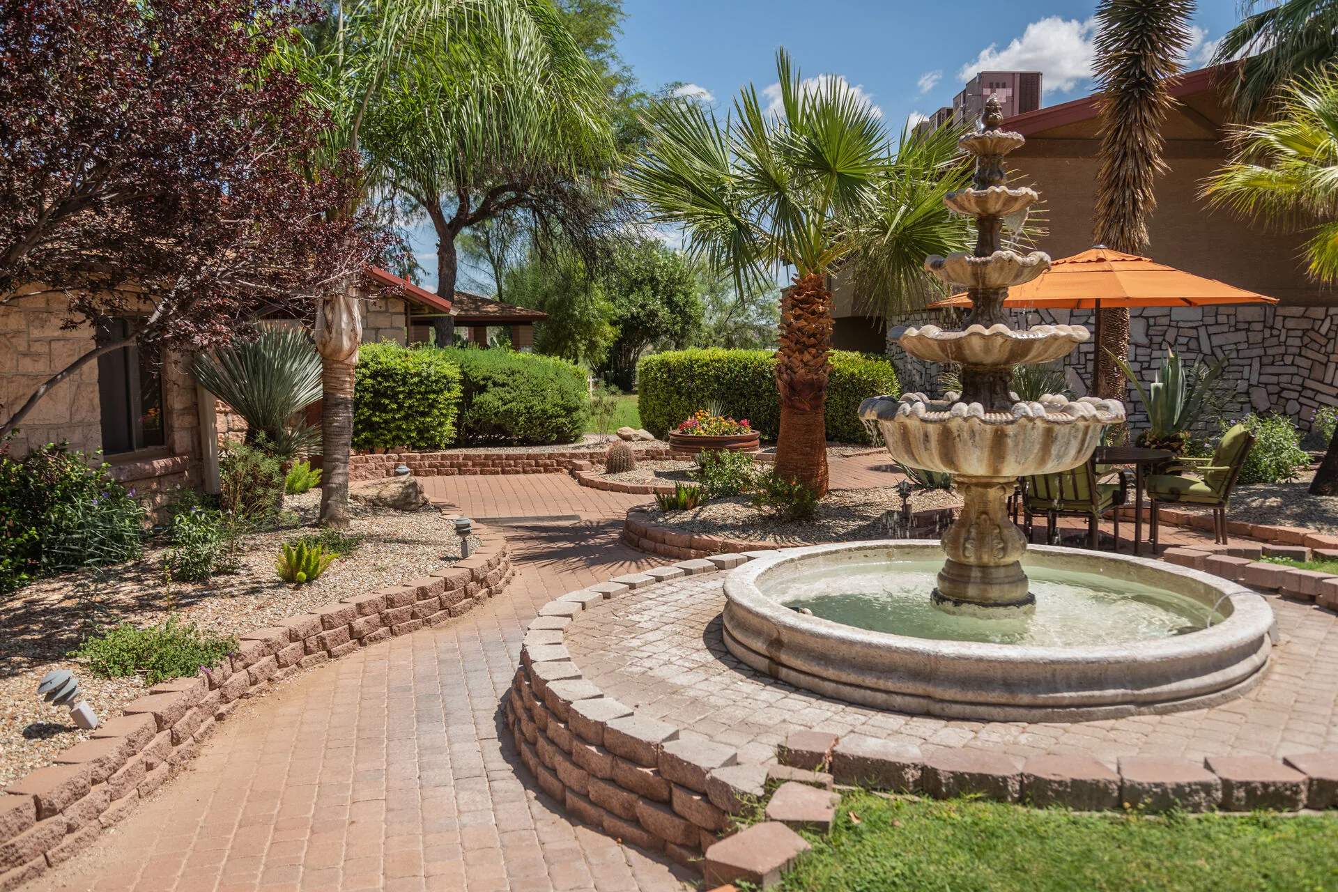 Brick-paved courtyard with a tiered stone fountain, palms and an orange parasol beside campus buildings
