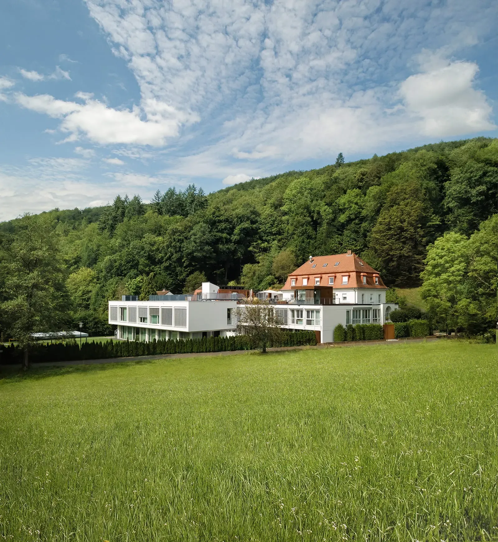 Red-roofed villa and white modern wing behind a green meadow, with forested hills under a cloudy blue sky