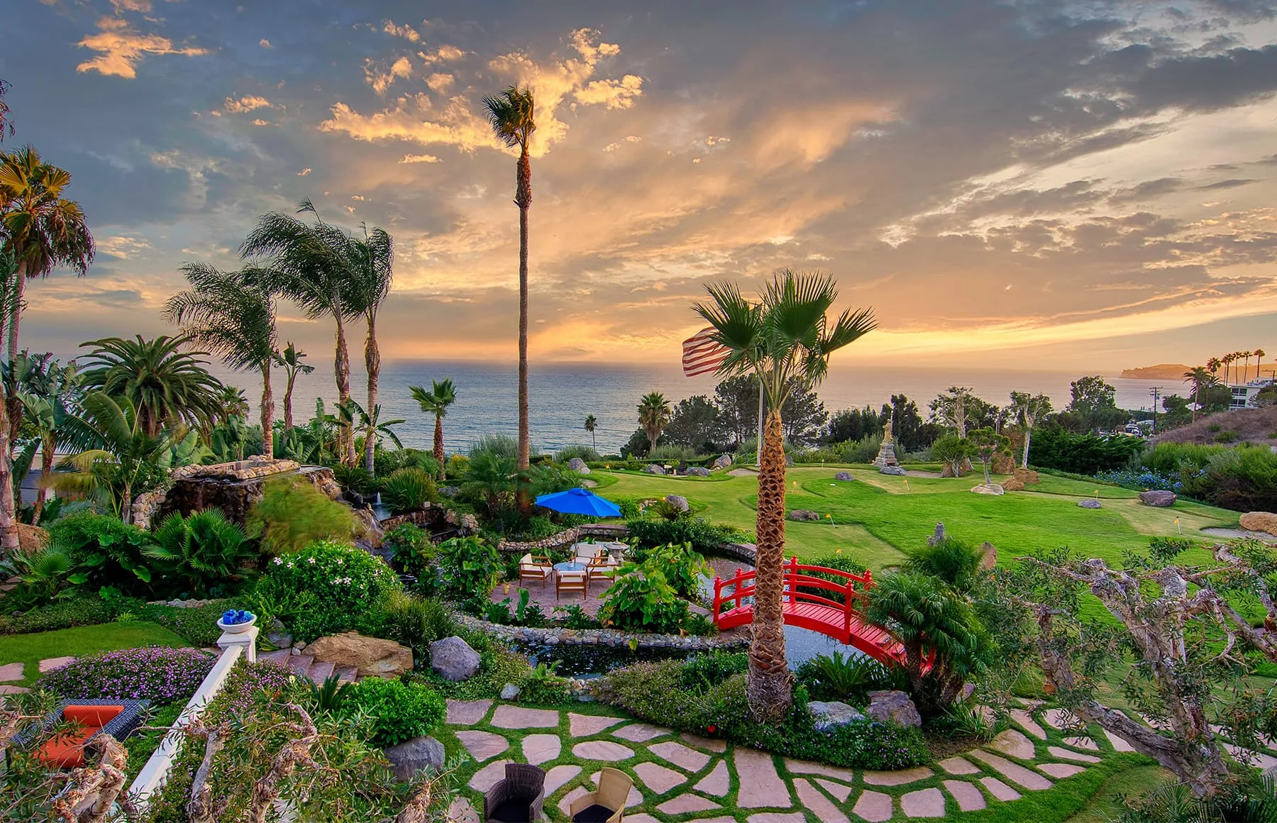 Terraced gardens with palms, stone paths and a red footbridge overlooking the ocean at sunset