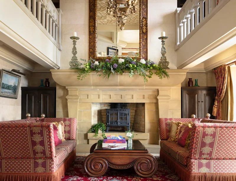 Hall with a carved stone fireplace, two patterned red sofas, a gilded mirror and a gallery above