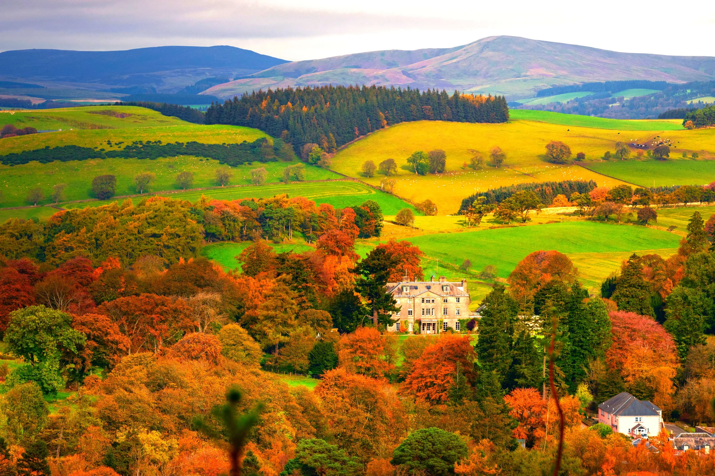 Autumn woodland in red and orange around the house, with green and yellow fields and hills beyond