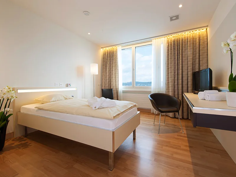 Bright private room with a single bed, wooden floor, desk, armchair and long curtained windows