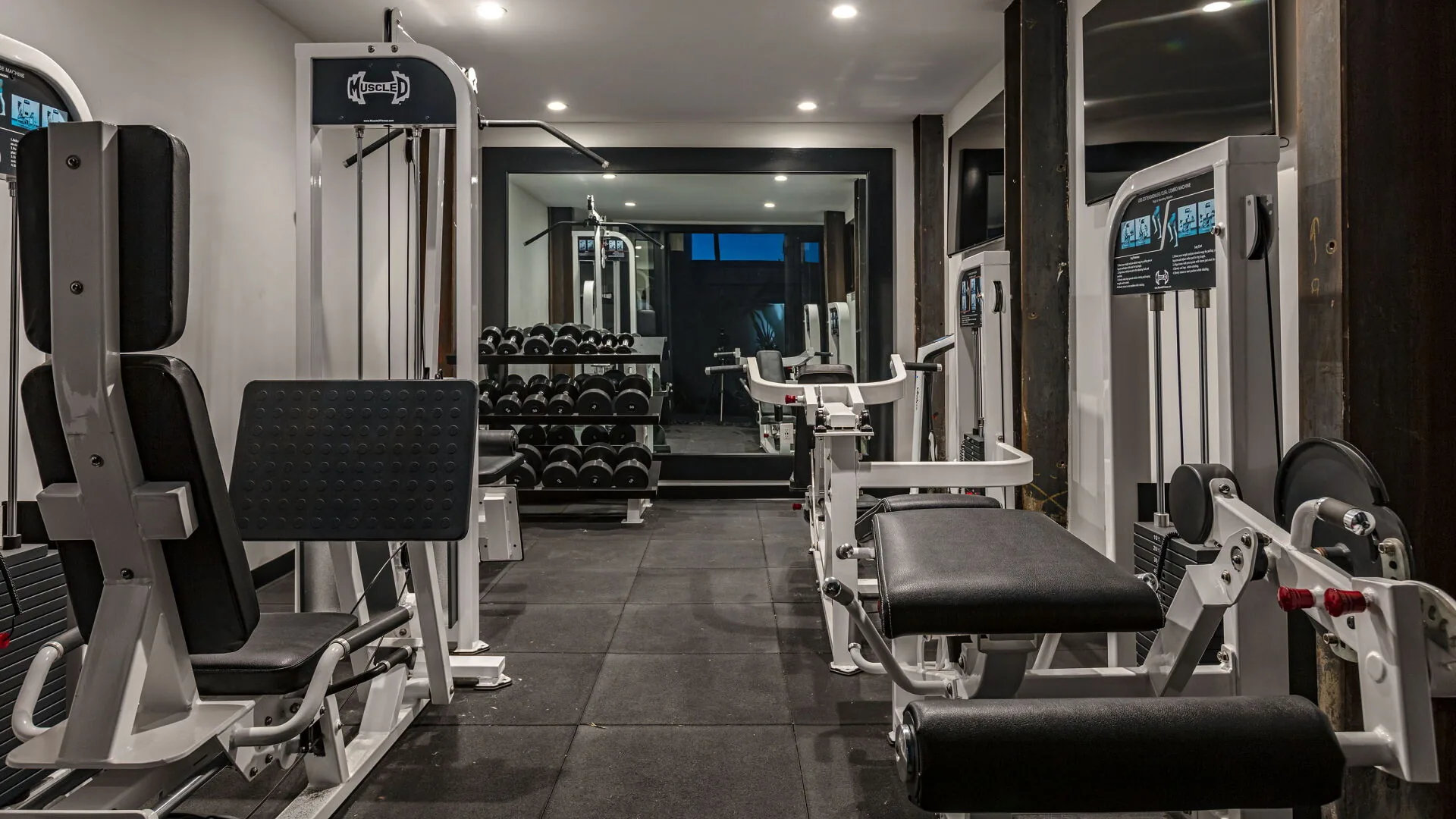 Home gym with weight machines, dumbbell rack and a mirrored wall on a dark tiled floor