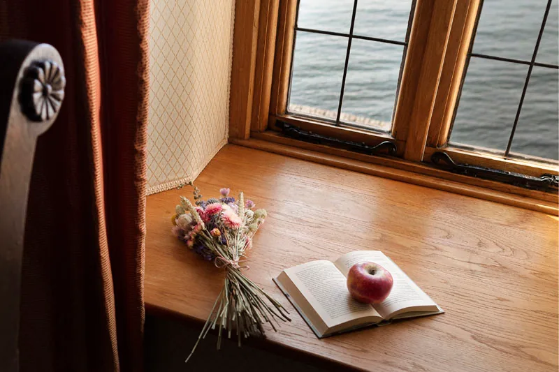 Wooden window seat with an open book, an apple and wild flowers below leaded windows facing water