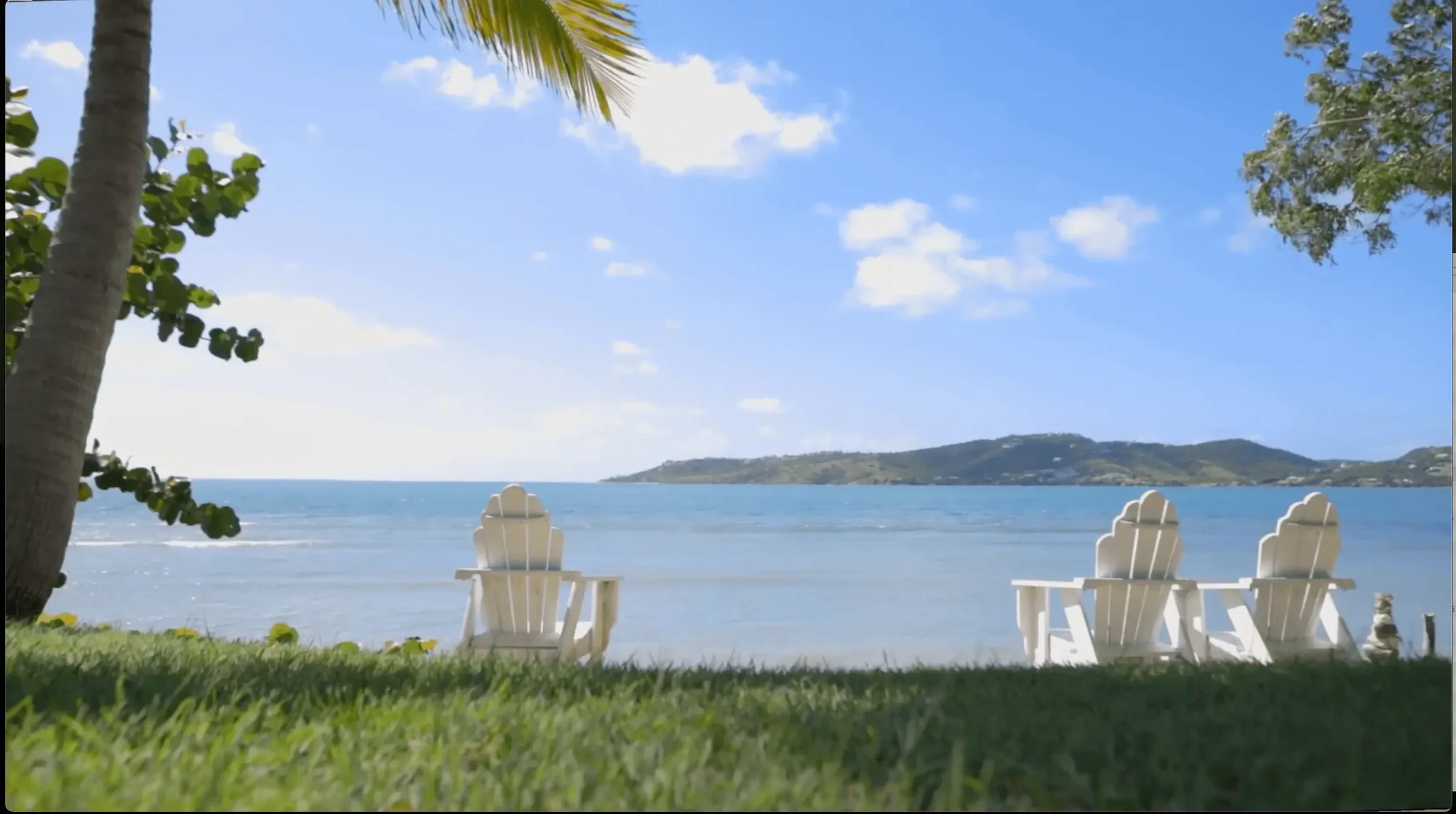 White Adirondack chairs on a seaside lawn looking across a calm bay to a hilly island under blue sky