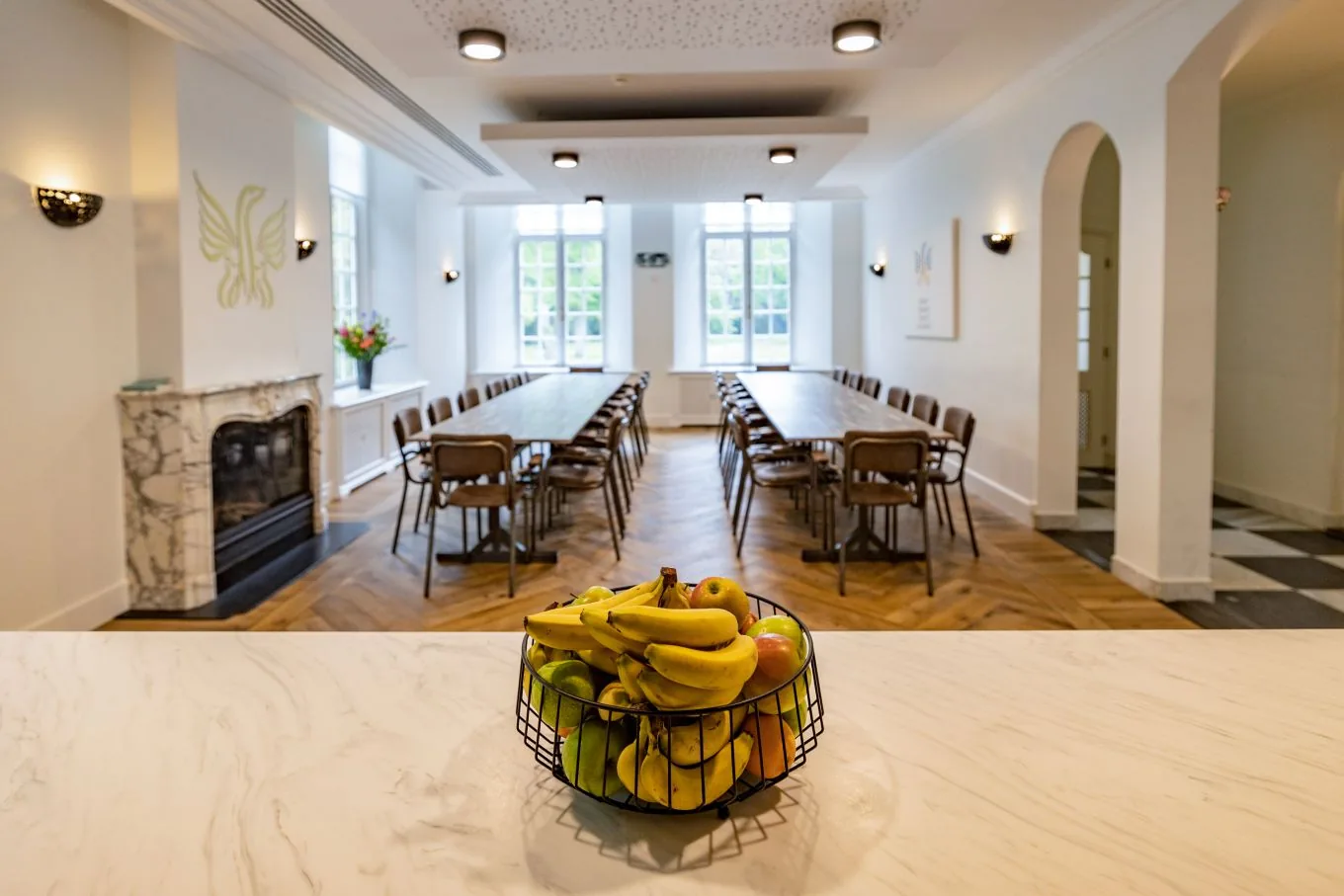 Long dining room with rows of tables and chairs, a marble fireplace and a bowl of bananas in front