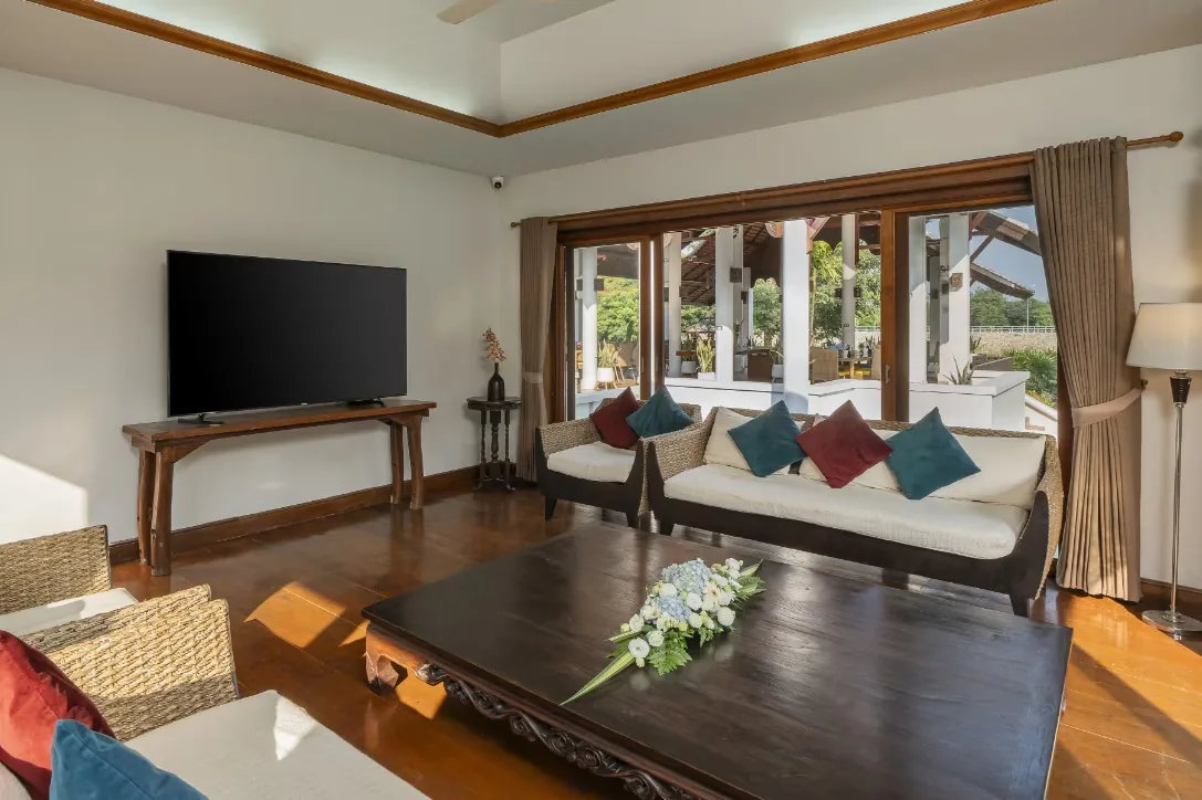 Lounge room with a cream sofa, coloured cushions, a wooden coffee table and a television by large windows