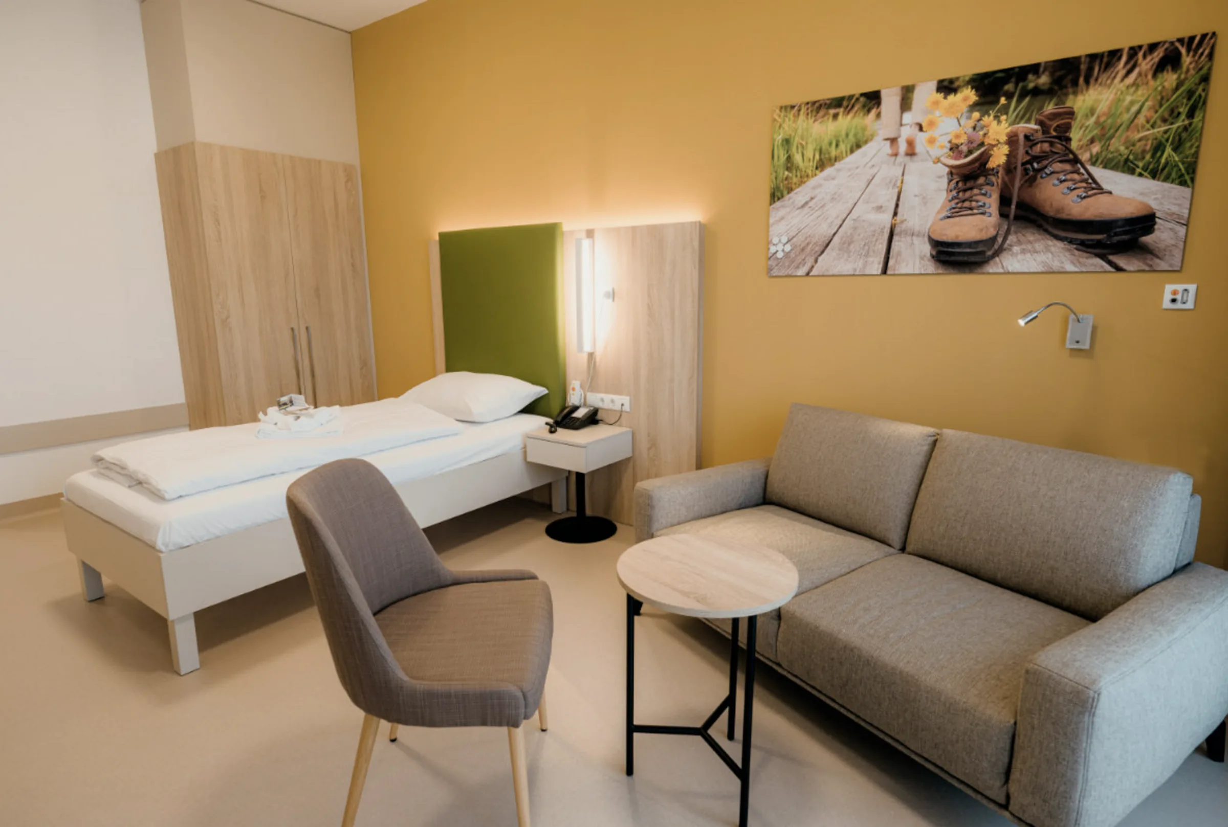 Single patient room with bed, grey sofa, round side table and a wall print showing hiking boots