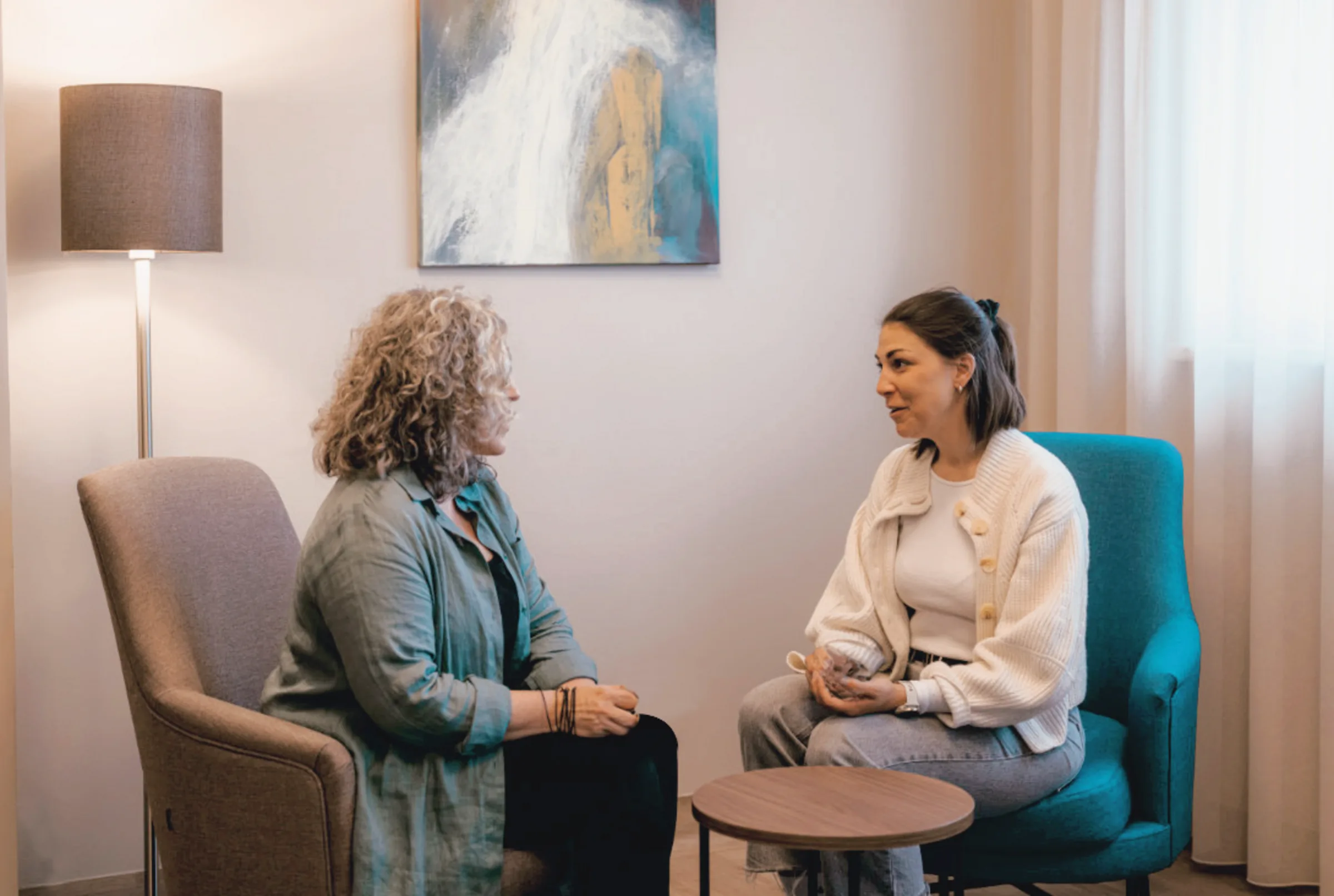 Two women talking in armchairs in a bright consultation room with a floor lamp and abstract painting