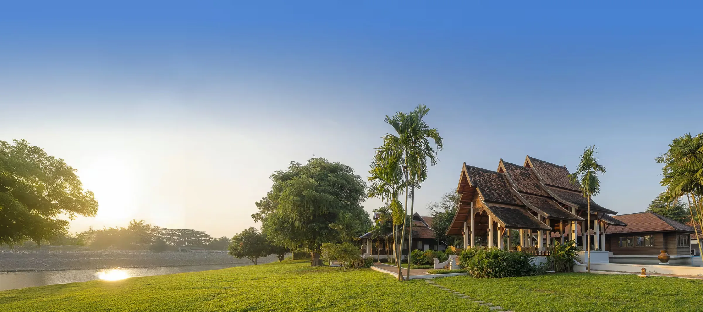 Northern Thai style pavilion with tiered roofs beside a river and palm trees on a lawn at sunset