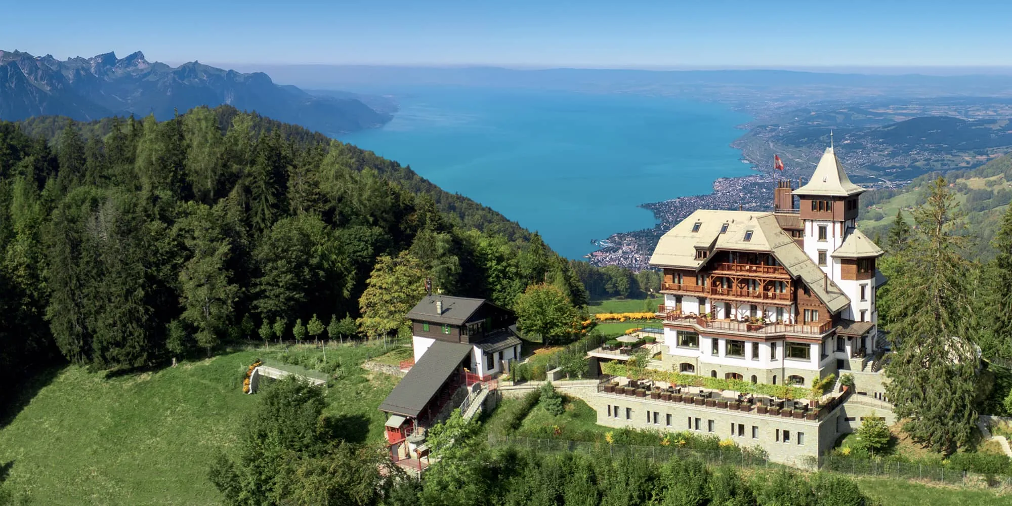 Aerial view of the chateau style clinic on a green slope above Lake Geneva with mountains beyond