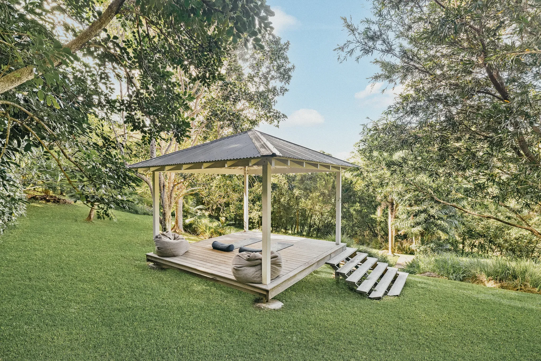 Open timber gazebo with a cushioned daybed on a green lawn surrounded by tall trees