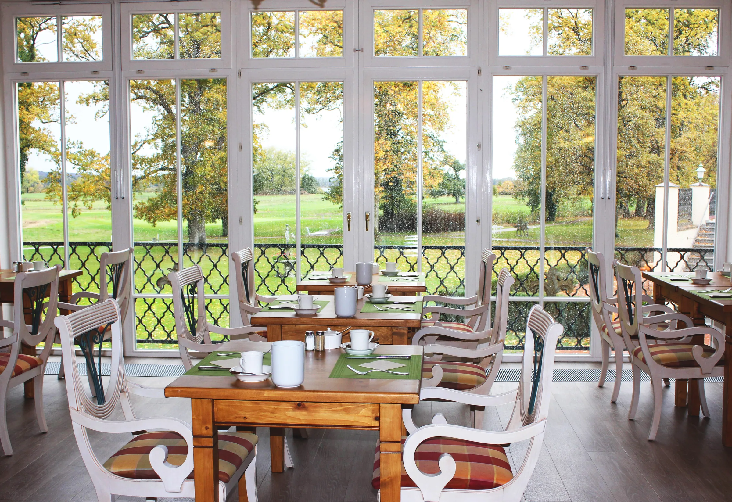 Breakfast room with wooden tables and white carved chairs before tall windows facing the park