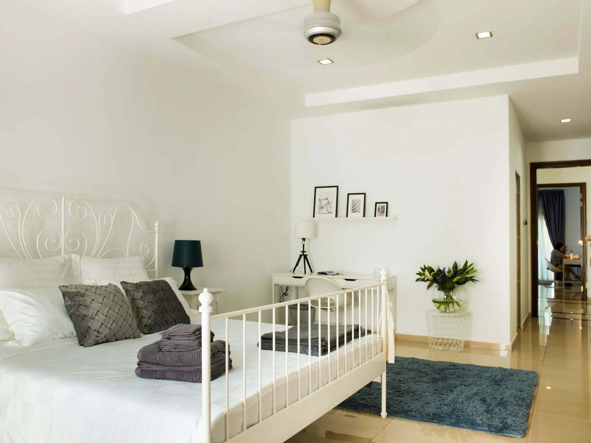 Bright single bedroom with a white iron bed, grey cushions, a desk with a plant and a doorway to the en suite