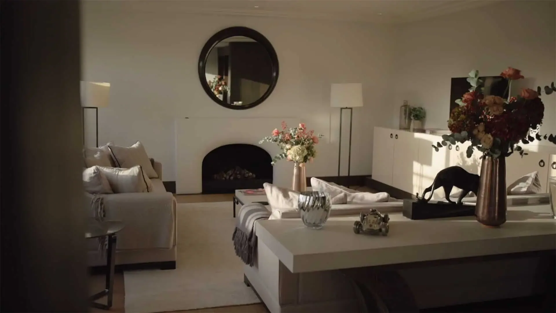 Dimly lit lounge with pale sofas, a lit fireplace, a round mirror and a vase of dried flowers