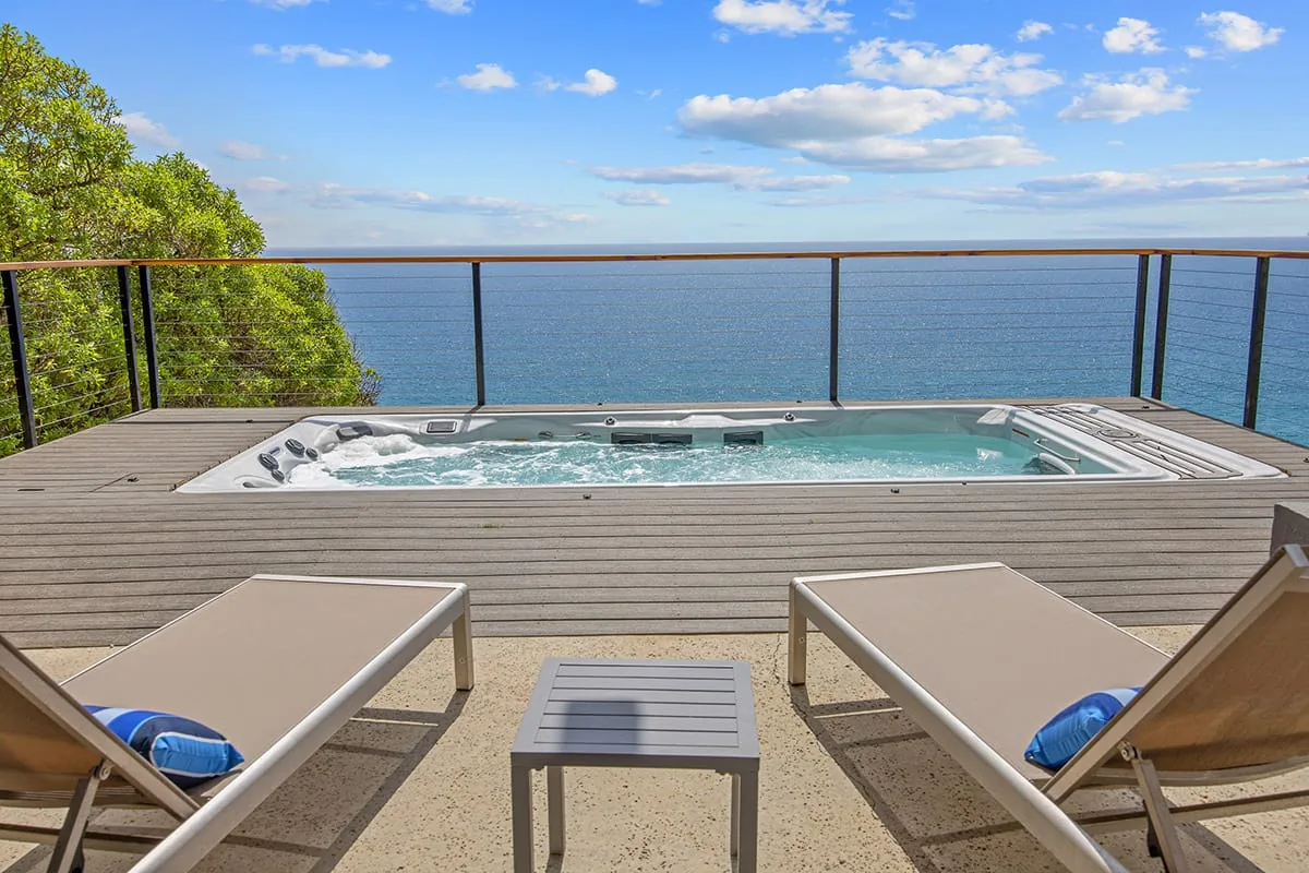 Sunken swim spa on a wooden deck with two lounge chairs and a glass railing above the Pacific