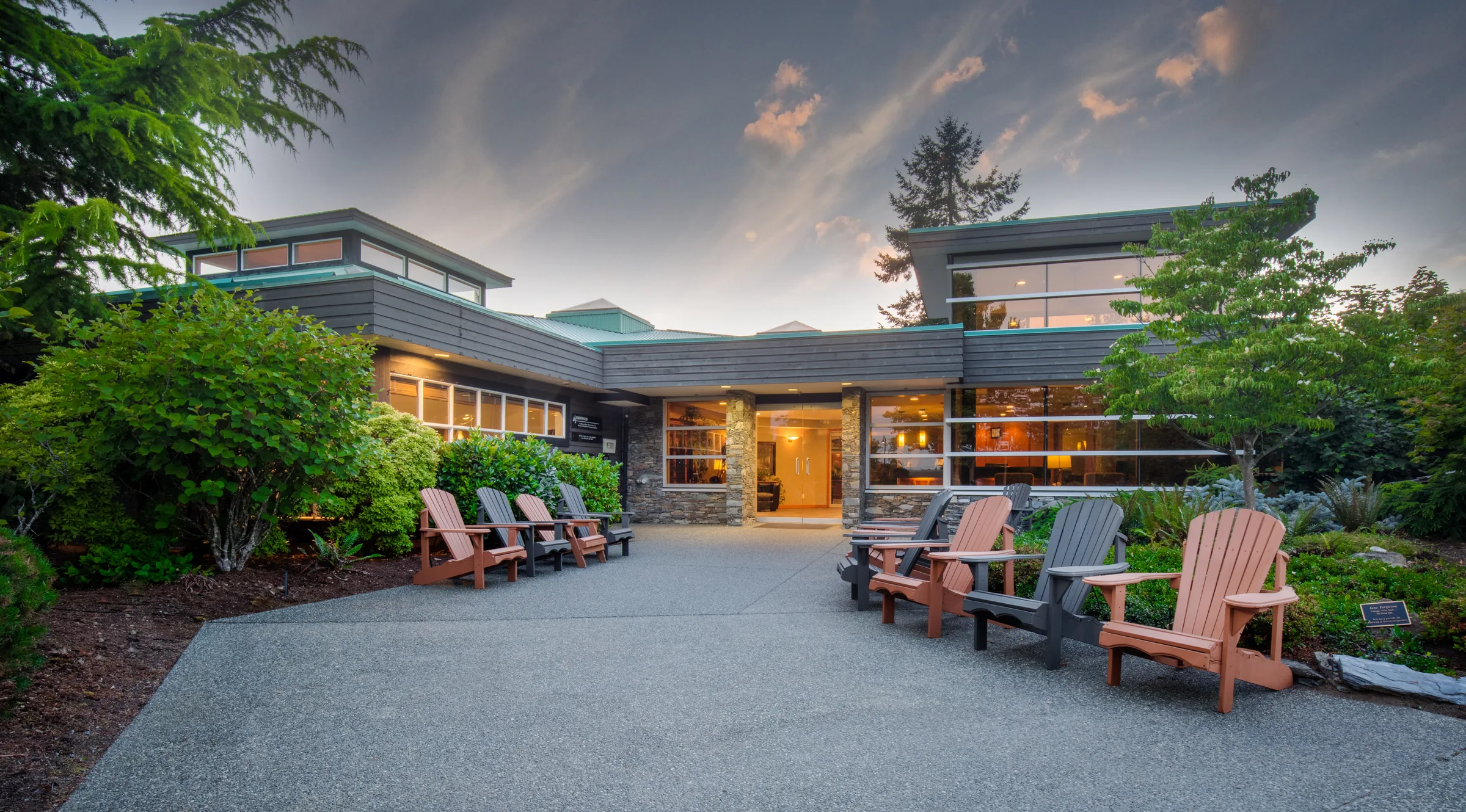 Lit front entrance of the low timber and stone building at dusk with Adirondack chairs on the forecourt