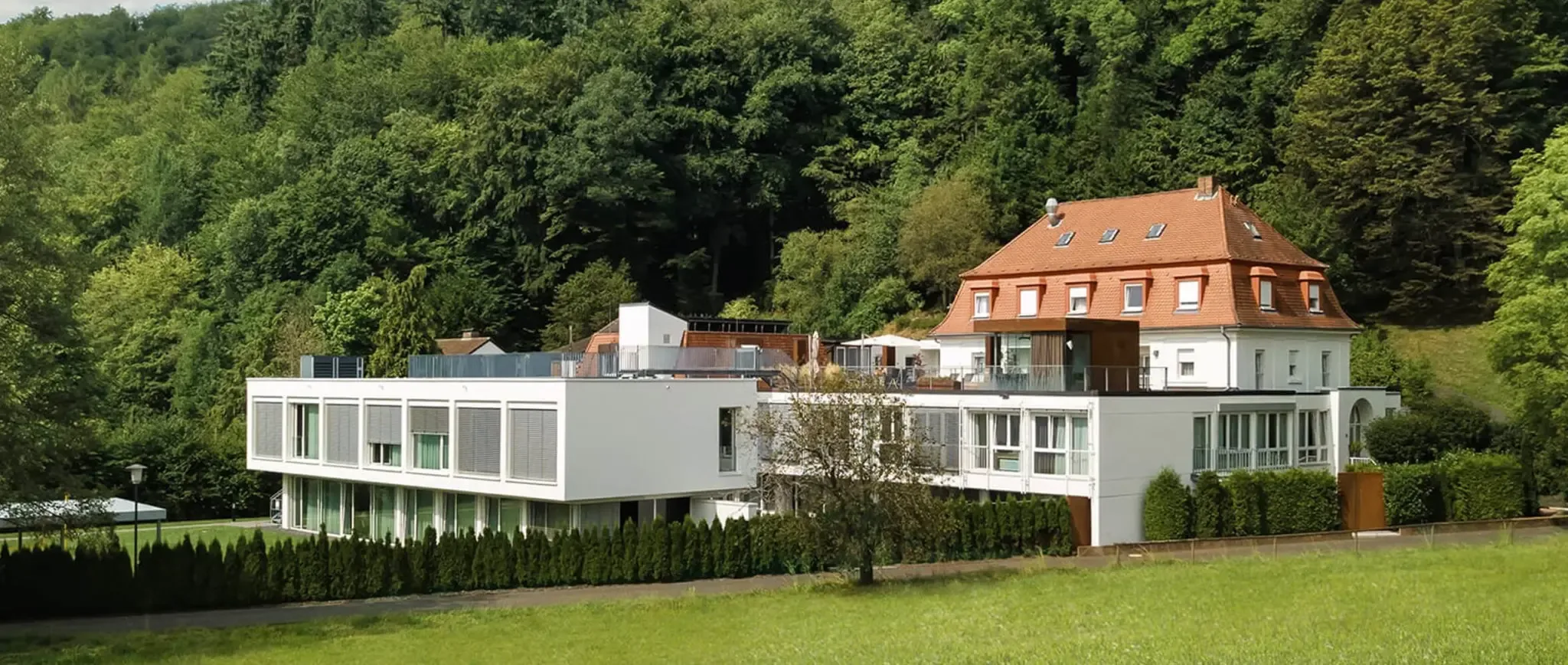 Wide view of the historic villa and flat-roofed modern wing set against a wooded hillside above a lawn